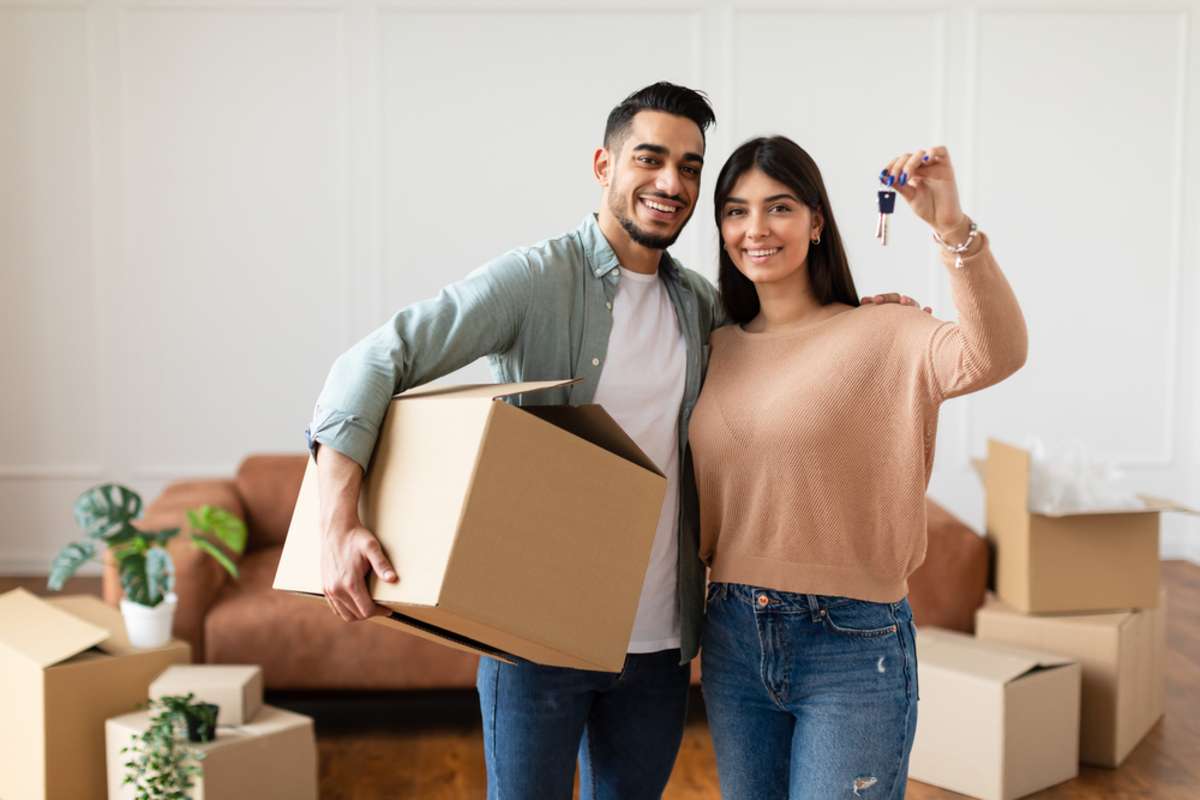 Young Couple Showing Keys And Holding Cardboard Box, Cheerful Guy And Lady Hugging After Moving In New Apartment