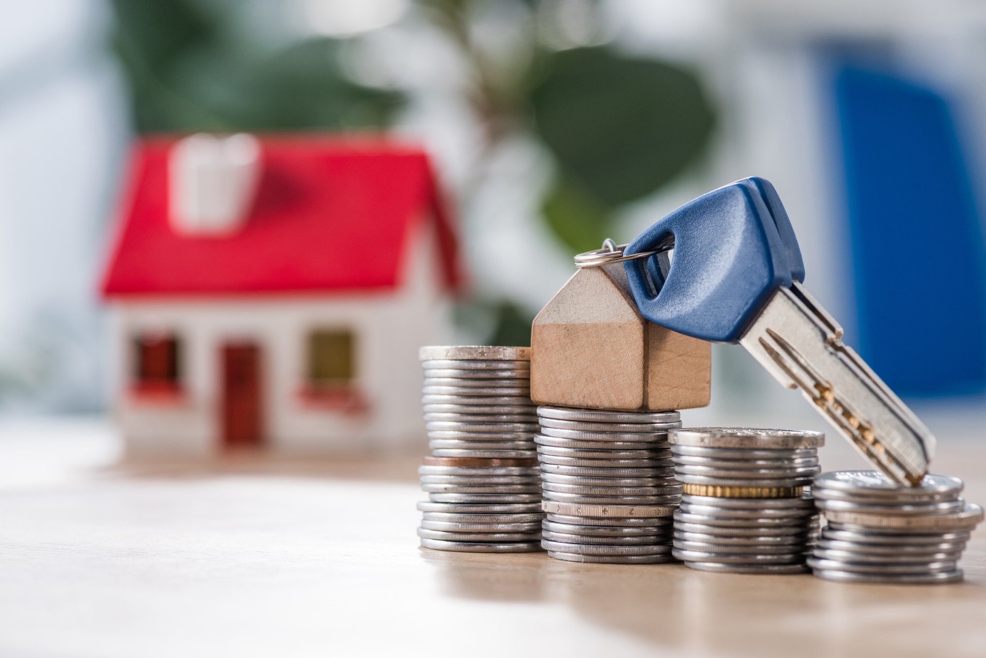 Selective focus of key with toy house trinket on stacks of coins near house model on wooden table