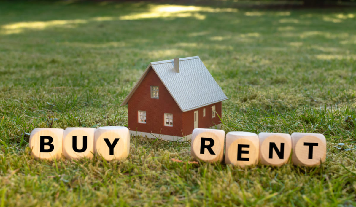 A miniature house placed on green meadow with wooden letter blocks that say BUY and RENT.